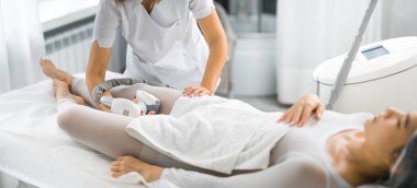 20 'li yaşlarının sonundaki çekici Asyalı kadın, endermoloji ekipmanlarıyla profesyonel Kafkas kozmetikçisi tarafından bacaklarındaki lipomasaj sırasında sırt üstü yatıyordu. Yüksek kalite fotoğraf