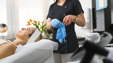 Siyah üniformalı tanınmayan beyaz kozmetikçi, güzel beyaz kadın müşterisinin SPA tedavisine hazırlanmak için mavi koruyucu eldivenler giyiyor. Yüksek kalite fotoğraf