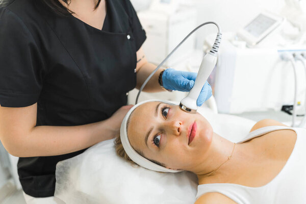 Indoor closeup trait of attractive blonde adult caucasian woman in white tank top during microdermabrasion procedure performed by unrecognizable beautician in black uniform. High quality photo