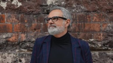 Gri saçlı, sakallı, gözlüklü ve ceketli orta yaşlı bir adam yıpranmış tuğla duvarın önünde dikilip kameraya gülümsüyor. Yatay açık hava videosu. Yüksek kalite 4k görüntü