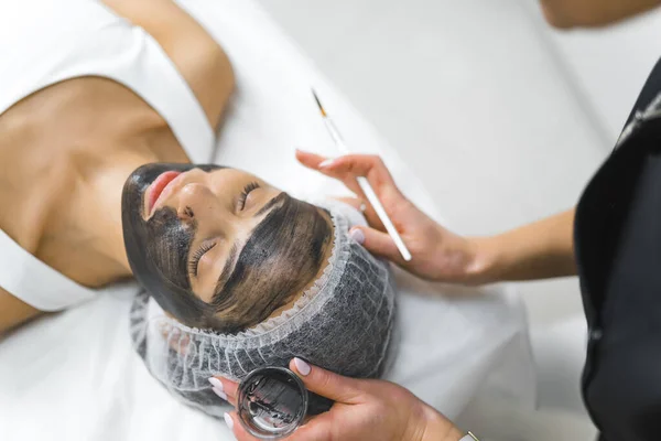 Professional facial procedure in SPA salon. Caucasian unrecognizable beautician applying black coal mask onto the face of beautiful young Latin girl. Relaxation concept. High quality photo