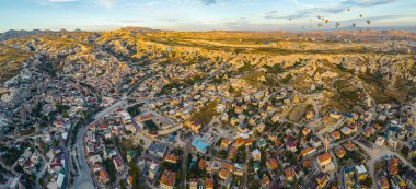 Kapadokya, Türkiye 'nin kayalık tepeleri, dağları ve arka planda uçan sıcak hava balonları ile panoramik hava manzarası. Parlak sarı güneş ışığı. Turistik bir yer. Yatay çekim. Yüksek kalite