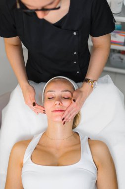 Geleneksel Japon yüz gerdirme masajı sırasında beyaz kolsuz tişörtlü, Avrupalı orta yaşlı bir kadının dikey yüksek açılı görüntüsü. Estetisyen POV. Yüksek kalite fotoğraf