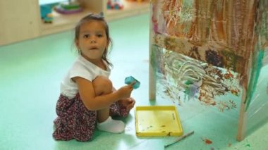 Koyu saçlı küçük bir kız anaokulunda yerde oturup selofan kutusuna resim yapıyor. Anaokulunda yaratıcı aktiviteler yapıyor. Yüksek kalite 4k görüntü