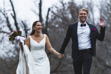Yeni evliler mutlu bir şekilde koşup gülüyorlar. Açık havada sonbahar foto çekimleri. Kolsuz gelinlik giyen Türk gelin zarif İskandinav kocasıyla el ele tutuşuyor. Yüksek kalite fotoğraf