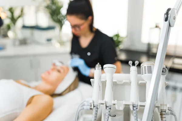 Various cosmetic devices used for different types of facials. Focus on the foreground. Unrecognizable caucasian female beautician doing microdermabrasion procedure on her clients face - blurred in