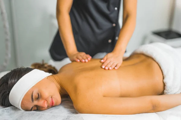 Taking care of ones health through SPA body care. Satisfied happy caucasian young woman with hairband above her forehead during relaxing back massage performed by unrecognizable massage therapist in