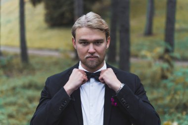 Yakışıklı, sarışın, sakallı, İskandinav damadı 20 'li yaşlarının sonunda siyah kelebek kravatını düzeltiyor ve kameraya bakıyor. Arka planda bulanık orman. Yüksek kalite fotoğraf