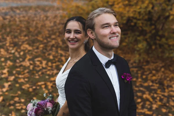 stock image Happiest moment of life - wedding day with best friend. Dreamy blond bearded caucasian man in black tuxedo with butterfly tie standing in front of his smiling Turkish bride. Golden leaves on the grass