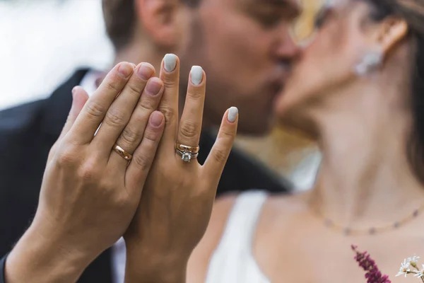 Concept of wedding and jewelry. Hands of two ethnic people with golden shiny wedding rings on ring fingers in the foreground. Defocused pair of heterosexual spouses kissing in the background. High