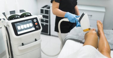 Lazer epilasyon konsepti. Kafkas lazer temizleme uzmanı siyah üniformalı profesyonel lazeri beyaz müşterisinin bacaklarının yanında tutuyor. Yüksek kalite fotoğraf