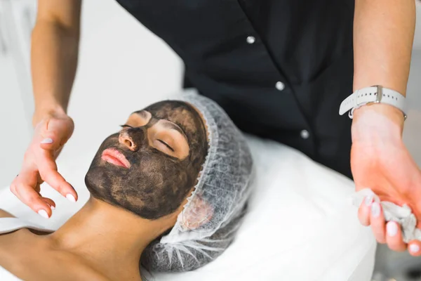 Relaxed Latin woman in her 30s lying down on SPA bed in protective hair cap when unrecognizable dermatologist in black uniform spreads charcoal mask on her face. High quality photo