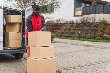 Ağır işçilik, paket teslimatı. Tam boy açık hava çekimi. Dost canlısı siyah kurye, karton paketler için depo vagonunu kullanırken kameraya bakıyor. Kutularla dolu beyaz bir minibüs.