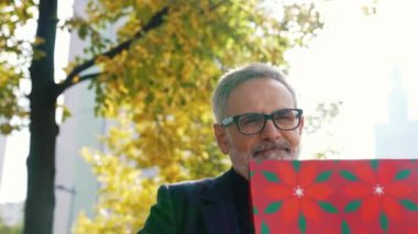 Gülümseyen, pozitif, sakallı, gözlüklü gri saçlı bir adam kırmızı bir hediye çantası tutuyor ve parktaki birine veriyor. Yüksek kalite 4k görüntü