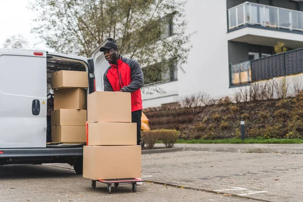 stock image Full-length outdoor shot of African-American delivery man using hand truck to deliver cardboard boxes. White delivery van with opened trunk full of packages. Modern building block in the background