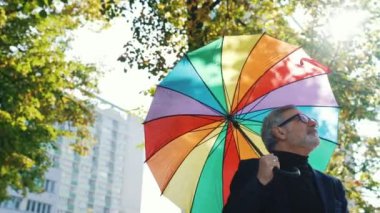 Rahat, orta yaşlı, sakallı, beyaz saçlı, spor ceketli ve balıkçı yakalı bir adam. Kafasını parlak renkli bir şemsiyeyle örtüyor, güneşli parkta yürüyor. Yüksek