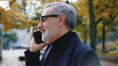 Yakışıklı, sakallı, takım elbiseli, gri saçlı adam telefonda arkadaşıyla konuşuyor. Parkta yapraklar döküldü. Yüksek kalite 4k görüntü