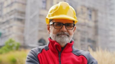 Gülümseyen, kendine güvenen, gri saçlı, üniformalı, gözlük takan, inşaat alanının önünde kameraya bakan orta yaşlı profesyonel bir işçinin portresi. Yüksek