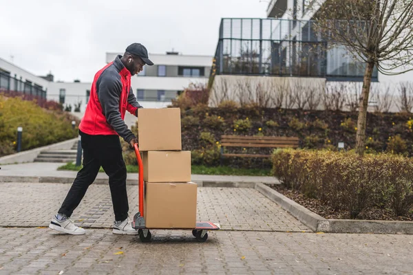 Black young adult delivery guy wearing work uniform walking on sidewalk pushing trolley with stacked cardboard box packages. Horizontal outdoor shot. High quality photo
