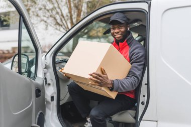 Kırmızı kazaklı, siyah şapkalı genç bir teslimatçı elinde paketle beyaz minibüsün kapısında oturmuş gülümseyerek kameraya bakıyor. Yatay açık hava çekimi. Yüksek kalite fotoğraf