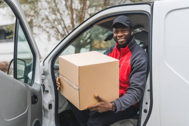 Dost canlısı, güler yüzlü, 20 'li yaşlarının ortasında, sürücü koltuğunda oturan, kapısı açık ve elinde karton bir paket tutan siyah bir adam. Kameraya bakıyorum. Pozitif titreşimler. Yüksek kalite fotoğraf