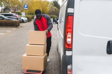 Siyah, genç, iş üniformalı bir teslimatçı beyaz minibüsten yük arabasına karton paketler boşaltıyor. Yatay açık hava çekimi. Yüksek kalite fotoğraf