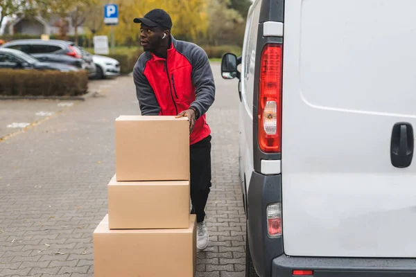 stock image Young adult black delivery man standing next to white van with unloaded parcels stacked on trolley. Horizontal outdoor shot. High quality photo