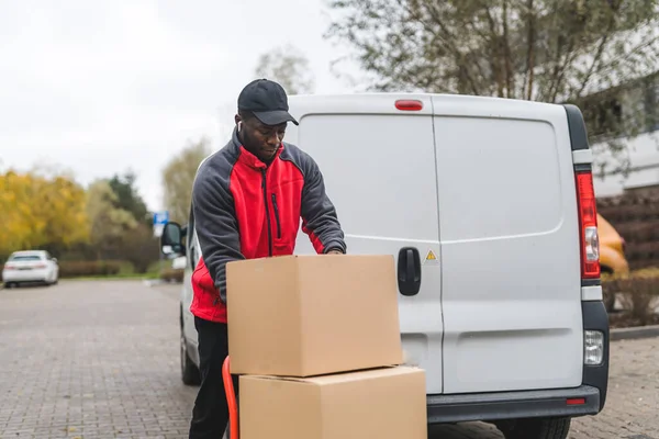 stock image Package delivery concept. Black handsome male courier using hand truck to deliver big packages packed in cardboard boxes. White delivery van in the background. High quality photo