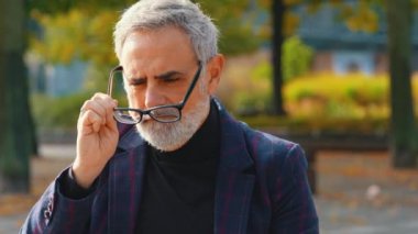 Şık, sakallı, takım elbiseli, gri saçlı gözlerini kısarak, parkta dururken daha iyi görebilmek için gözlüklerini takan bir adam. Yüksek kalite 4k görüntü