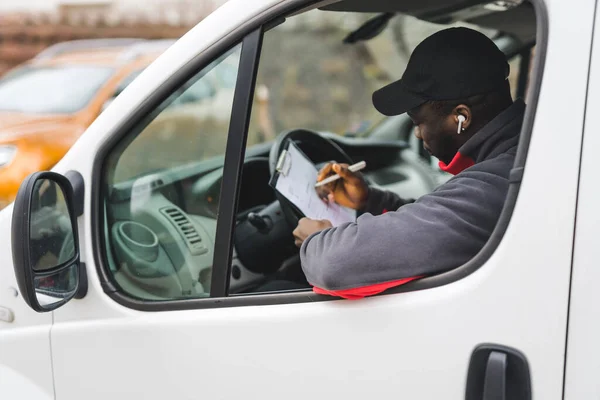 Black young adult delivery guy wearing work uniform and black cap sitting in driver seat of white van filling out socuments. Horizontal outdoor shot . High quality photo
