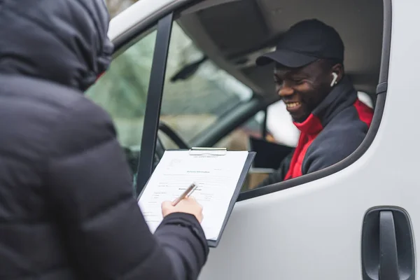 Black young adult delivery guy sitting in driver seat of white van smiling looking out the window at client signing papers. Horizontal shot . High quality photo