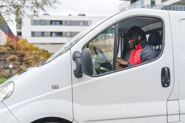 Black young adult delivery guy sitting inside white van working on laptop computer. Delivery service. Horizontal outdoor shot. High quality photo