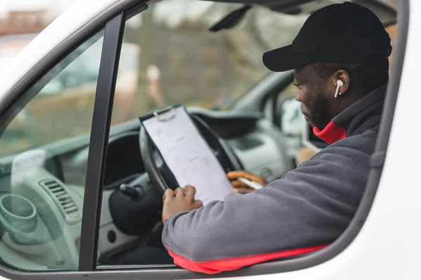 Close up of young adult black delivery guy wearing pullover and black cap sitting in driver seat of white van checking filled out documents. Horizontal shot. High quality photo