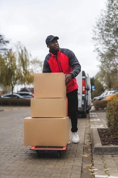 Young adult black delivery guy wearing red uniform pullover carrying parcels stacked on trolley. Vertical outdoor shot. High quality photo