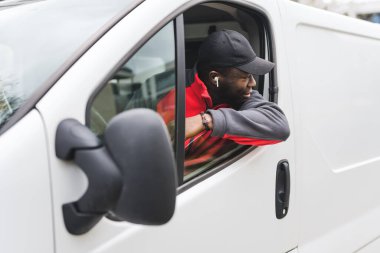 Arkasına bakmak için pencereden sarkan beyaz minibüs kullanan iş üniforması giyen genç siyahi bir teslimatçı. Yatay çekim. Yüksek kalite fotoğraf