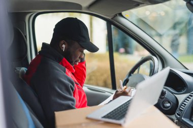 Minibüsün sürücü koltuğunda oturan genç zenci teslimatçı evrakları doldurup dizüstü bilgisayarla çalışıyor. Yatay çekim. Yüksek kalite fotoğraf