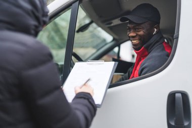 Genç, yetişkin, siyah bir adam, beyaz minibüsün içinde oturmuş, pencereden dışarı bakıyor ve müşterilerine evrakları imzalarken gülümsüyor. Yatay çekim. Yüksek kalite fotoğraf