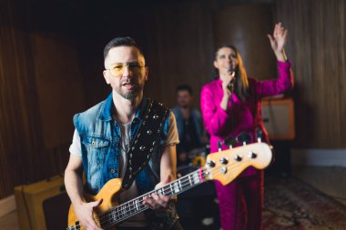 Sarı cam gözlüklü sakallı orta yaşlı adam elektro gitarla çalıyor. Arka planda bulanık kadın vokalist ve erkek davulcu var. İç mekan konser konsepti. Yüksek kalite fotoğraf
