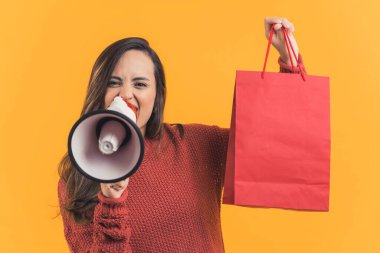 Kırmızı alışveriş çantasını elinde tutan Latin Amerikalı kadın megafonla heyecanlı bir ifadeyle konuşuyor. Reklamcılık. Satılık. Alışverişe. Sarı arka plan yatay stüdyo çekimi. Yüksek kalite fotoğraf