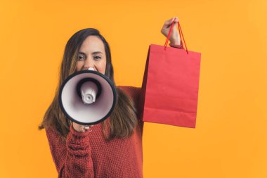 Latin Amerikalı bir kadın kameraya bakıp elinde kırmızı bir çantayla megafonla konuşuyor. Satılık. Alışverişe. Reklamlar. Sarı arka plan yatay stüdyo çekimi. Yüksek kalite fotoğraf