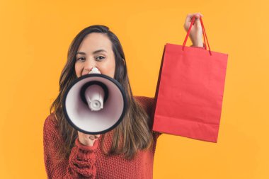 Kahverengi saçlı, elinde alışveriş çantası ve megafonla satış ve indirim ilan eden genç bir kadın. Stüdyoda orta boy turuncu bir fotoğraf çekildi. Yüksek kalite fotoğraf