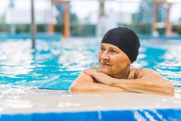 Pensive senior caucasian grandmother in black hair cap relaxing at the side of indoor swimming pool. Active pensioners. High quality photo