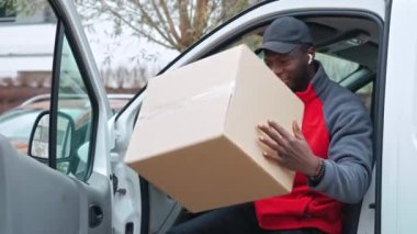 Siyah, yetişkin bir teslimatçı elinde karton kutuyla beyaz minibüsten gülümsüyor. - Evet. Yüksek kalite 4k görüntü