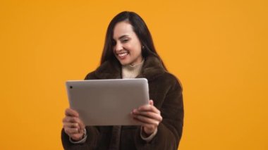 Kışlık ceketli neşeli Latin Amerikalı kadın elinde tablet, turuncu arka plan orta teknoloji konsepti. Yüksek kalite 4k görüntü