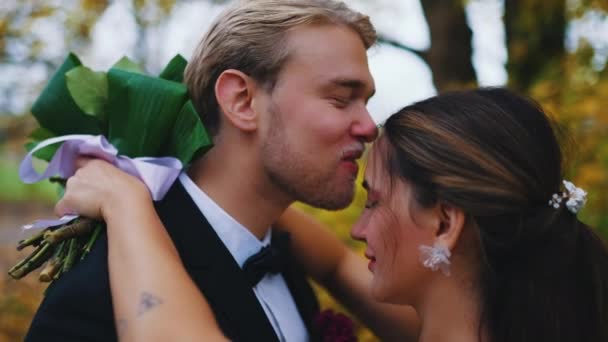 新婚旅行の間の素敵な優しいシーン 北欧の新郎の背中のビューは 彼の新婚トルコ人の妻の額にキスを与える 背景には緋色の秋のオレンジ色の葉 屋外写真撮影 — ストック動画