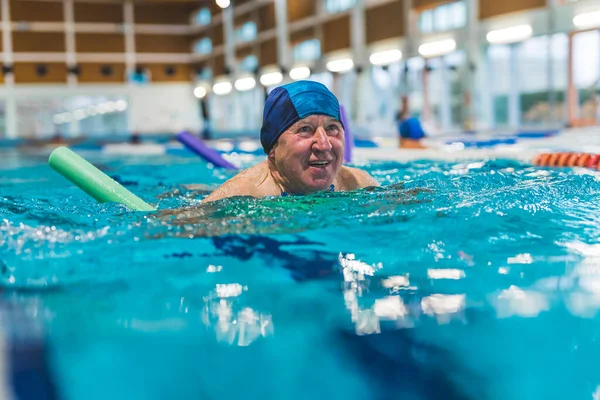 Stock image Portrait of positive smiling caucasian senior adult man in dark blue head cap swimming in a sport pool with the use of foam green pool noodle. High quality photo