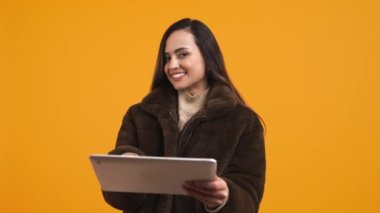 Kışlık ceketli mutlu Latin Amerikalı kadın elinde tablet, turuncu arka plan orta teknoloji ve iş kadını konseptiyle izole edilmiş. Yüksek kalite 4k görüntü