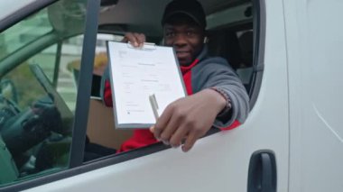 Beyaz minibüsün sürücü koltuğunda oturan genç siyahi teslimatçı müvekkilinin imzalaması için pencereden kağıtları uzatıyor. - Evet. Yüksek kalite 4k görüntü