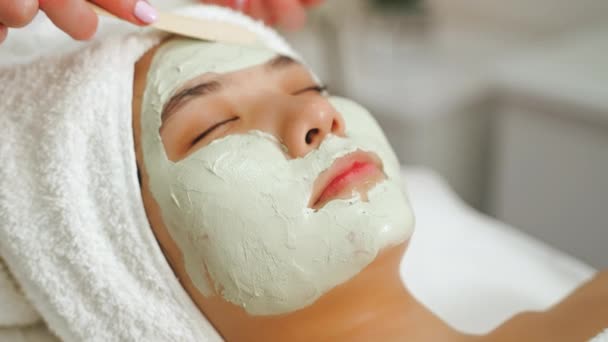 Spa的深层净化程序 一位20多岁 面带微笑 头戴白毛巾的漂亮亚洲女人的画像躺在Spa床上 而经验丰富的美容师则戴上了粘土面罩 — 图库视频影像