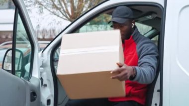Kırmızı kazaklı, siyah şapkalı genç bir teslimatçı elinde paketle beyaz minibüsün kapısında oturmuş gülümseyerek kameraya bakıyor. Yüksek kalite 4k görüntü